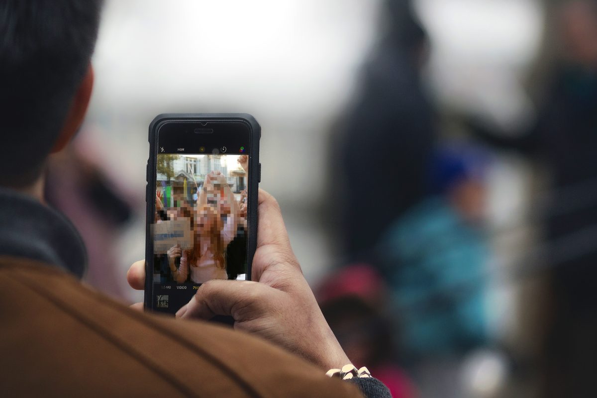 Une personne vue de dos filme avec son téléphone ; à l’écran, une manifestation où les visages sont pixelisés